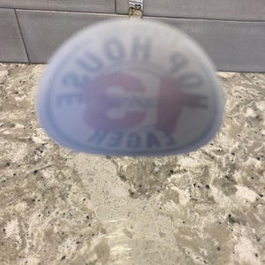 May include: A clear glass with a blurred logo that reads "HOP HOUSE LAGER". A measuring tape is visible in the background, showing approximately 3 inches. The glass sits on a speckled countertop.