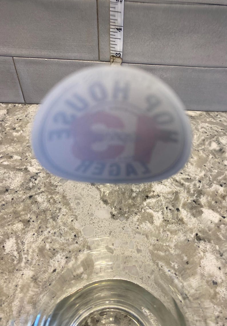 May include: A clear glass with a blurred logo that reads "HOP HOUSE LAGER". A measuring tape is visible in the background, showing approximately 3 inches. The glass sits on a speckled countertop.