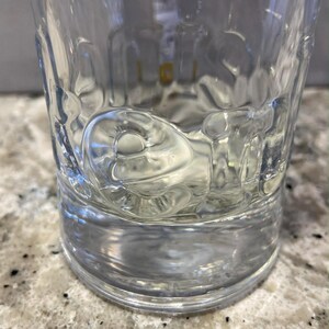 以下が含まれることがあります： 厚いベースの透明な模様入りのガラスタンブラー。ガラスの側面にはテクスチャード加工が施されており、光と影の遊びを作り出しています。タンブラーは空で、まだらの表面に置かれています。