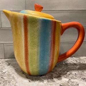 May include: A colourful ceramic jug with a striped design in shades of yellow, orange, red, green, blue, and white. The jug has a lid with a small orange knob and a handle.