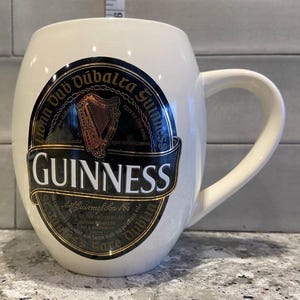 May include: White ceramic mug with a black and gold Guinness logo. The logo features the Guinness harp and the text "honn Oub Dubalra Guinnes" and "GUINNESS".