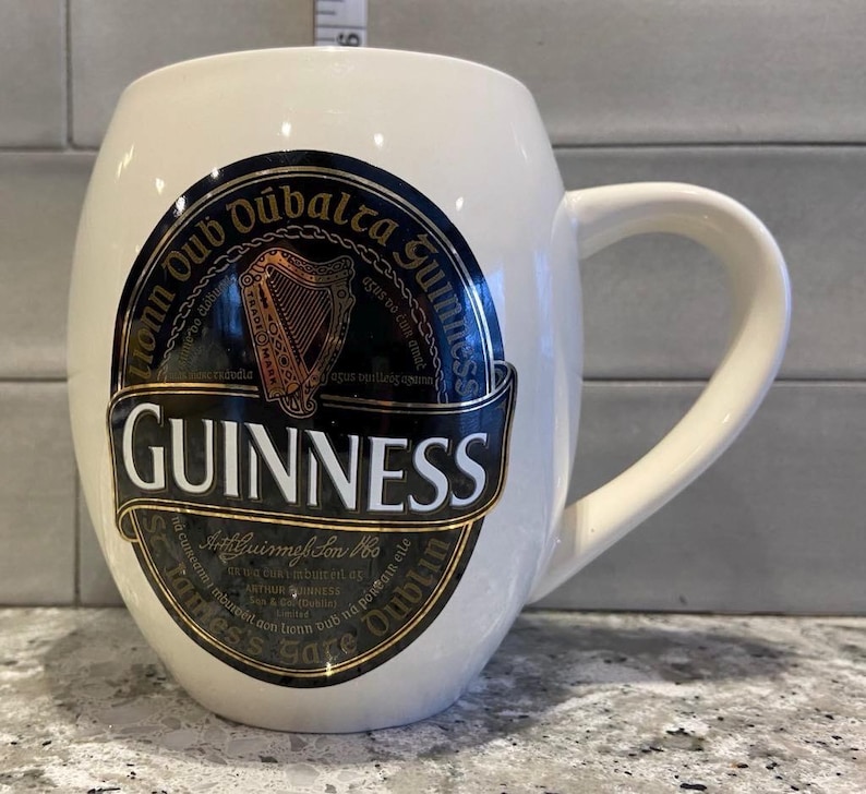 May include: White ceramic mug with a black and gold Guinness logo. The logo features the Guinness harp and the text "honn Oub Dubalra Guinnes" and "GUINNESS".