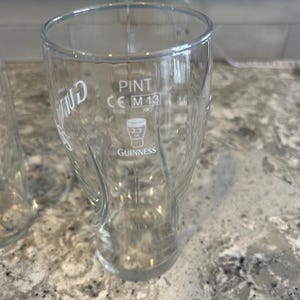 K&ouml;nnte beinhalten: Ein klares Pint-Glas mit dem Guinness-Logo und -Text. Das Glas hat ein geschwungenes Design und ist mit "PINT" und "2.3 UK UNITS" gekennzeichnet. Das Glas ist leer und steht auf einer gesprenkelten Arbeitsplatte.