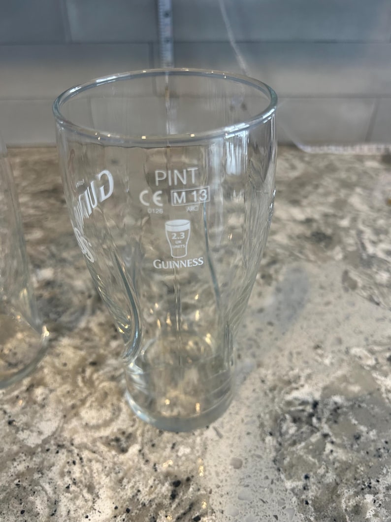 K&ouml;nnte beinhalten: Ein klares Pint-Glas mit dem Guinness-Logo und -Text. Das Glas hat ein geschwungenes Design und ist mit "PINT" und "2.3 UK UNITS" gekennzeichnet. Das Glas ist leer und steht auf einer gesprenkelten Arbeitsplatte.