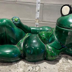 May include: A ceramic frog figurine in a reclining pose, painted in glossy emerald green. The frog wears black-framed glasses with white lenses. The figurine measures approximately 5 inches in length.