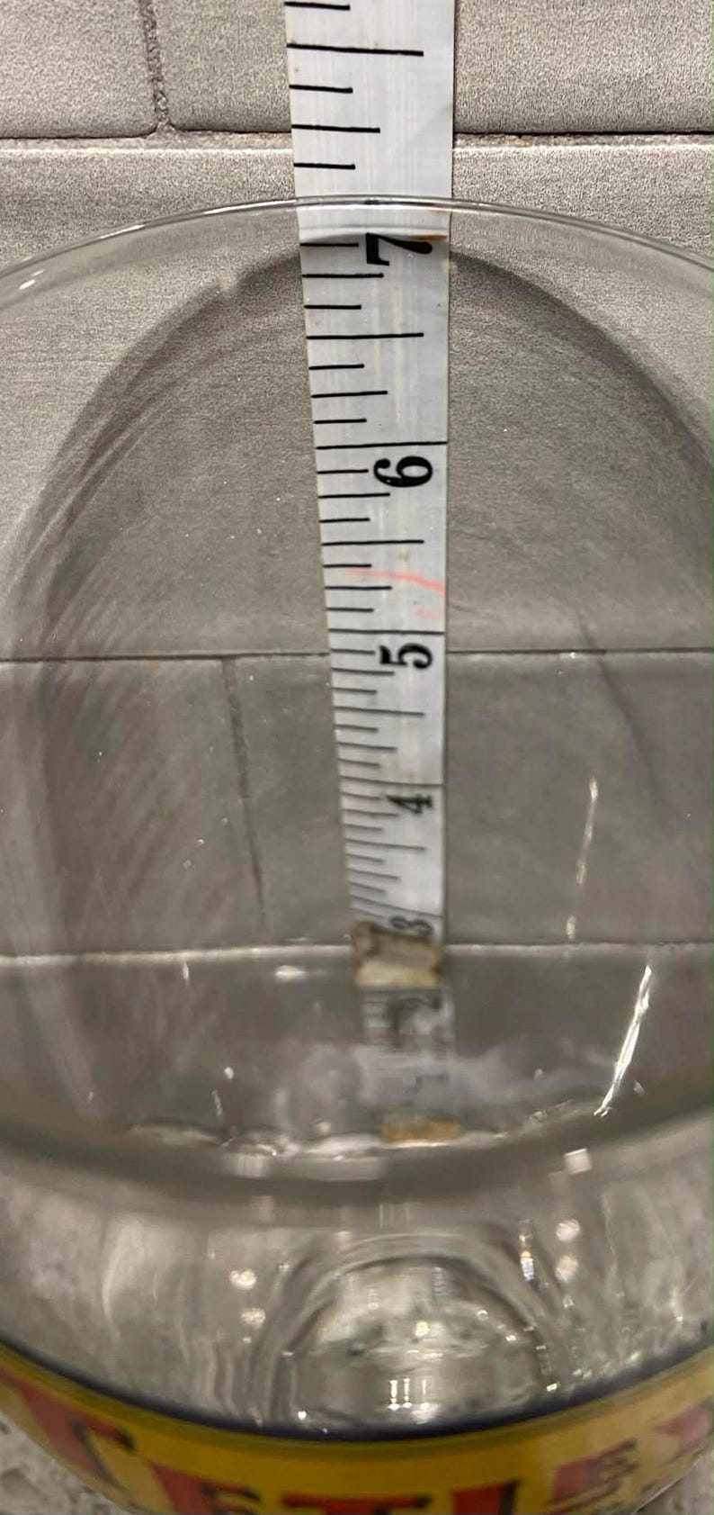 May include: A clear glass container with a measuring tape inside, showing a liquid level at approximately 3 inches. The tape is marked in inches, with clear black lines and numbers.