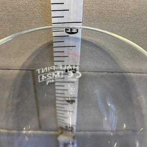 May include: A clear glass with a white measuring tape inside. The tape shows measurements in inches, with the top reading approximately 17.78 cm. The glass has a wide opening and a rounded base.