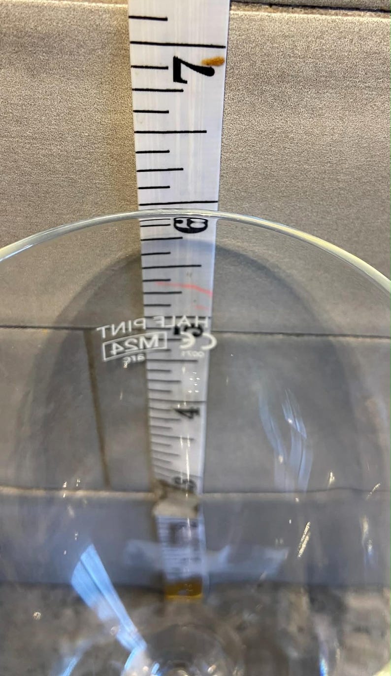 May include: A clear glass with a white measuring tape inside. The tape shows measurements in inches, with the top reading approximately 17.78 cm. The glass has a wide opening and a rounded base.