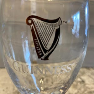 May include: A clear Guinness pint glass featuring the Guinness logo and the word "GUINNESS" in white. The logo is a black harp with the text "ESTD 1759". The glass is clean and ready for a pint.