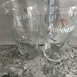 K&ouml;nnte beinhalten: Zwei transparente Guinness-Biergl&auml;ser mit dem Guinness-Logo und Harfendesign. Die Gl&auml;ser sind etwa 15 cm hoch. Die Gl&auml;ser stehen auf einer gesprenkelten grauen Oberfl&auml;che.