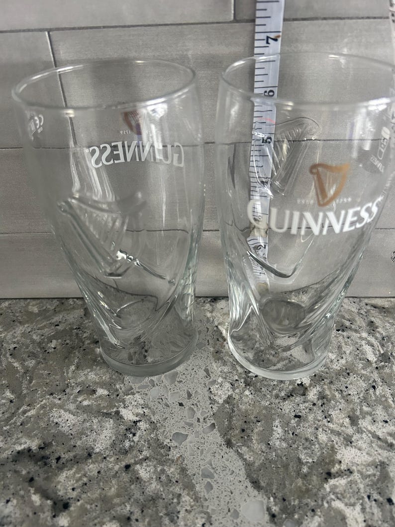K&ouml;nnte beinhalten: Zwei transparente Guinness-Biergl&auml;ser mit dem Guinness-Logo und Harfendesign. Die Gl&auml;ser sind etwa 15 cm hoch. Die Gl&auml;ser stehen auf einer gesprenkelten grauen Oberfl&auml;che.