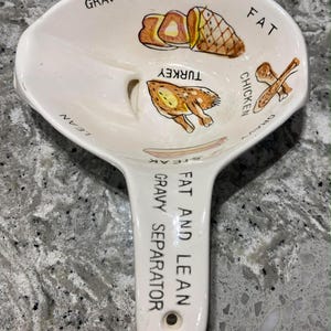 Puede incluir: Separador de salsa de cerámica blanca con asa. El cuenco presenta ilustraciones de jamón, pavo y pollo, con etiquetas de texto para "Gravy", "Fat" y "Lean". El asa dice "Fat and Lean Gravy Separator".