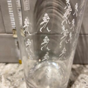 May include: A clear glass tumbler with a repeating white graphic of a striding gentleman. The glass is cylindrical and appears to be empty. The background is out of focus.