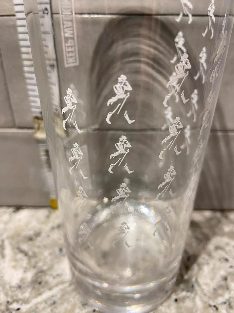May include: A clear glass tumbler with a repeating white graphic of a striding gentleman. The glass is cylindrical and appears to be empty. The background is out of focus.