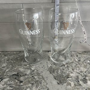 K&ouml;nnte beinhalten: Zwei transparente Guinness-Biergl&auml;ser mit dem Guinness-Logo in Gold und Wei&szlig;. Die Gl&auml;ser sind etwa 18 cm hoch. Die Gl&auml;ser haben ein einzigartiges, geschwungenes Design und sind leer.