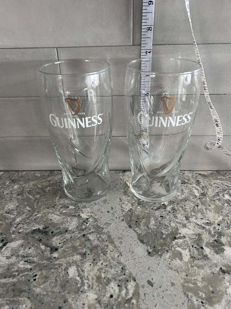 K&ouml;nnte beinhalten: Zwei transparente Guinness-Biergl&auml;ser mit dem Guinness-Logo in Gold und Wei&szlig;. Die Gl&auml;ser sind etwa 18 cm hoch. Die Gl&auml;ser haben ein einzigartiges, geschwungenes Design und sind leer.