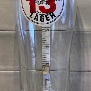 May include: A clear pint glass with a Guinness Hop House 13 Lager logo. A measuring tape is placed inside the glass, showing a height of approximately 5 inches.