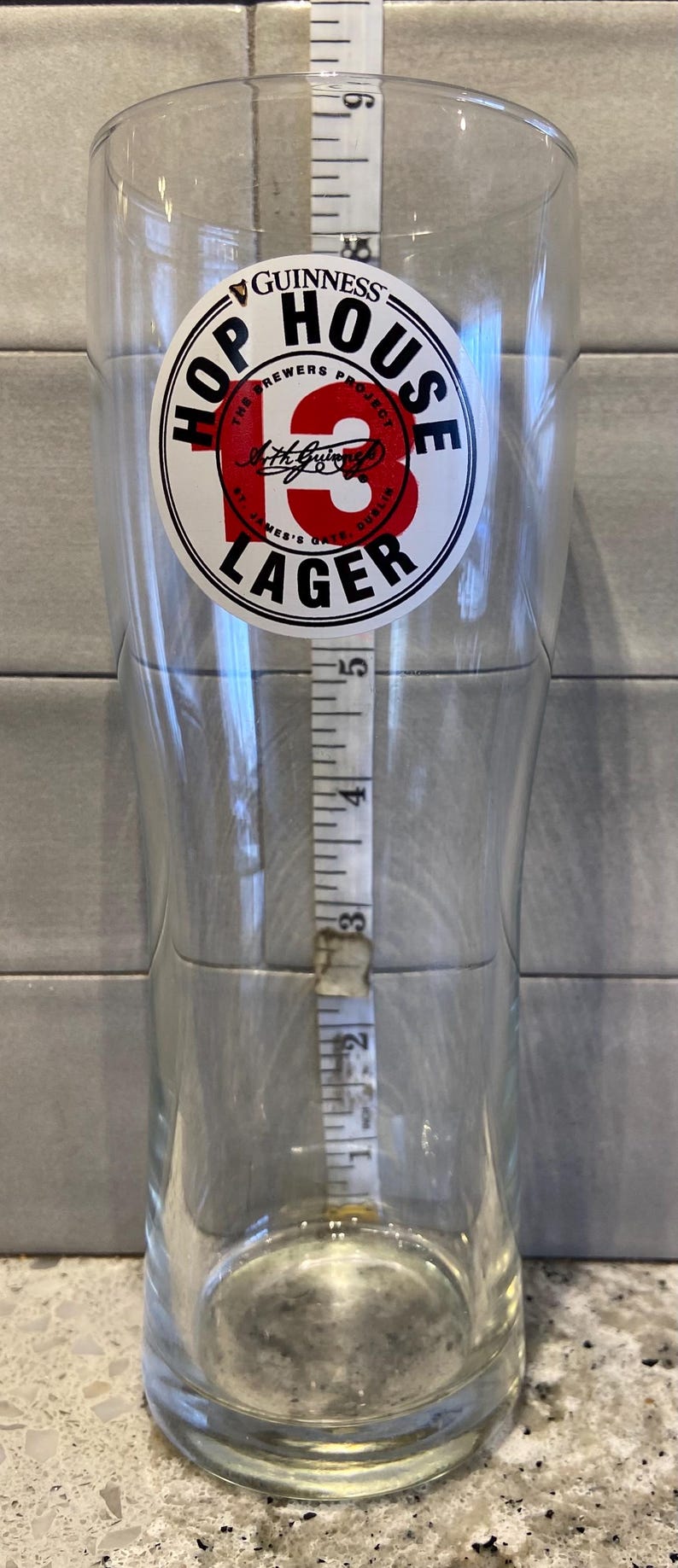 May include: A clear pint glass with a Guinness Hop House 13 Lager logo. A measuring tape is placed inside the glass, showing a height of approximately 5 inches.
