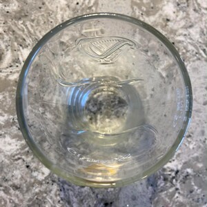 May include: Clear glass pint glass with the Guinness harp logo etched into the glass. The glass has a wide mouth and a slightly curved shape. The glass is empty and appears to be clean.