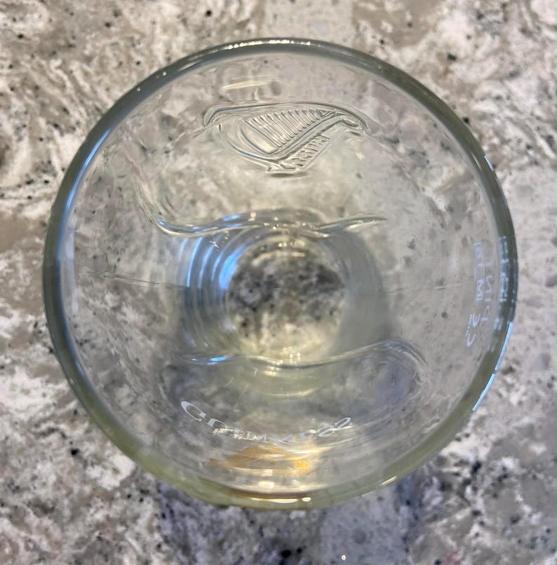 May include: Clear glass pint glass with the Guinness harp logo etched into the glass. The glass has a wide mouth and a slightly curved shape. The glass is empty and appears to be clean.