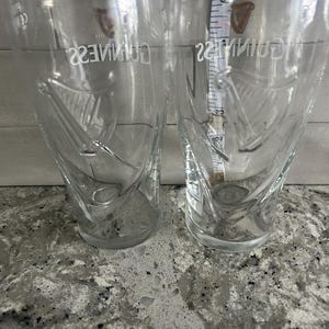 K&ouml;nnte beinhalten: Zwei transparente Biergl&auml;ser mit dem Guinness-Logo und Harfendesign. Die Gl&auml;ser sind leer und stehen auf einer gesprenkelten Arbeitsplatte. Ein Ma&szlig;band ist zwischen den Gl&auml;sern zu sehen.