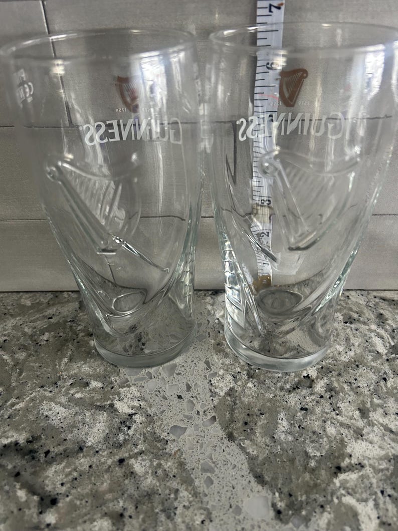 K&ouml;nnte beinhalten: Zwei transparente Biergl&auml;ser mit dem Guinness-Logo und Harfendesign. Die Gl&auml;ser sind leer und stehen auf einer gesprenkelten Arbeitsplatte. Ein Ma&szlig;band ist zwischen den Gl&auml;sern zu sehen.