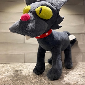 May include: A plush toy of a dark gray cat with large yellow eyes, a bright pink nose, and two white fangs protruding from its mouth. The cat wears a red collar with white dots and has a pointed, stylized head shape with tufted ears.
