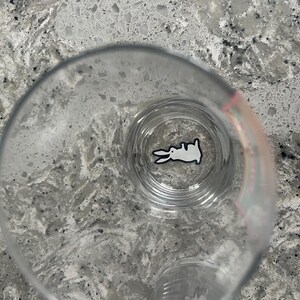 K&ouml;nnte beinhalten: Ein klares Glas mit einem wei&szlig;en Kaninchen-Design am Boden. Das Kaninchen ist in einem einfachen Cartoon-Stil mit schwarzen Umrissen dargestellt. Das Glas steht auf einer gesprenkelten, grauen Arbeitsplatte.