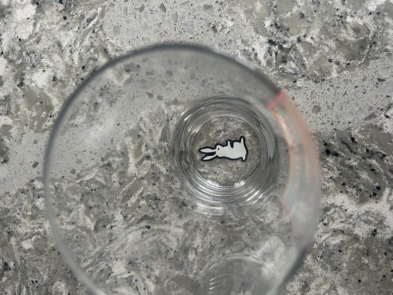 K&ouml;nnte beinhalten: Ein klares Glas mit einem wei&szlig;en Kaninchen-Design am Boden. Das Kaninchen ist in einem einfachen Cartoon-Stil mit schwarzen Umrissen dargestellt. Das Glas steht auf einer gesprenkelten, grauen Arbeitsplatte.