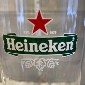 以下が含まれることがあります： ハイネケンのロゴが入った透明なグラス。ロゴは、白い「Heineken」の文字が入った緑色のバナーの上に赤い星が描かれています。バナーの下には白いホップの葉のデザインがあります。「EST. 1873」の文字も表示されています。