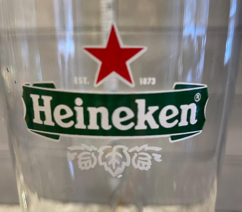 以下が含まれることがあります： ハイネケンのロゴが入った透明なグラス。ロゴは、白い「Heineken」の文字が入った緑色のバナーの上に赤い星が描かれています。バナーの下には白いホップの葉のデザインがあります。「EST. 1873」の文字も表示されています。