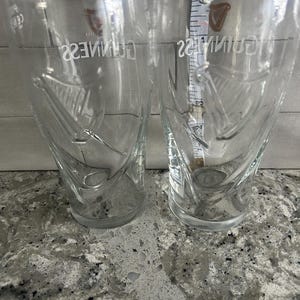 K&ouml;nnte beinhalten: Zwei transparente Guinness-Biergl&auml;ser mit dem Guinness-Logo und Harfendesign. Die Gl&auml;ser sind konisch und etwa 15 cm hoch. Sie stehen auf einer gesprenkelten grauen Oberfl&auml;che.