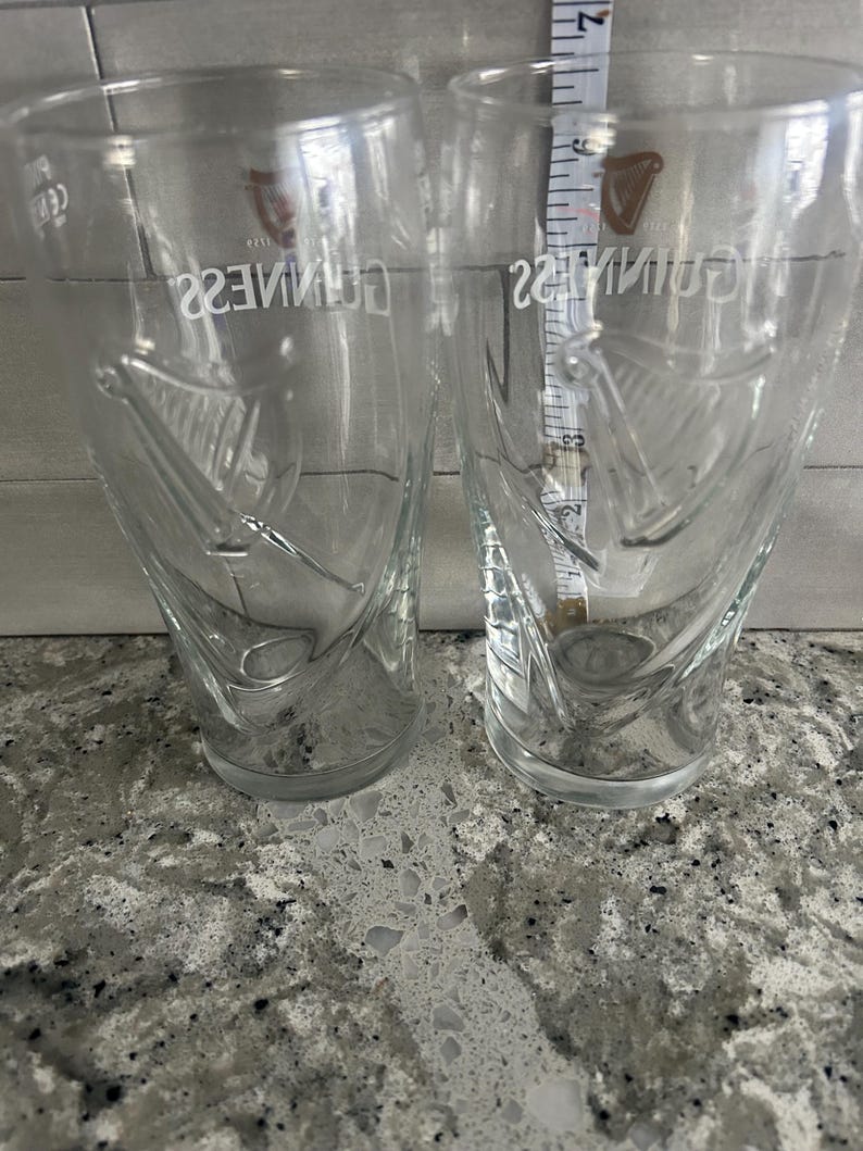 K&ouml;nnte beinhalten: Zwei transparente Guinness-Biergl&auml;ser mit dem Guinness-Logo und Harfendesign. Die Gl&auml;ser sind konisch und etwa 15 cm hoch. Sie stehen auf einer gesprenkelten grauen Oberfl&auml;che.