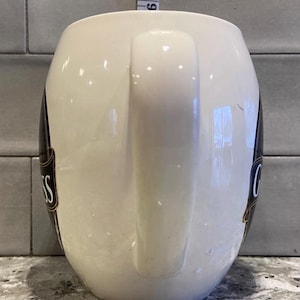 May include: White ceramic mug with a black and gold Guinness logo. The mug has a large handle.