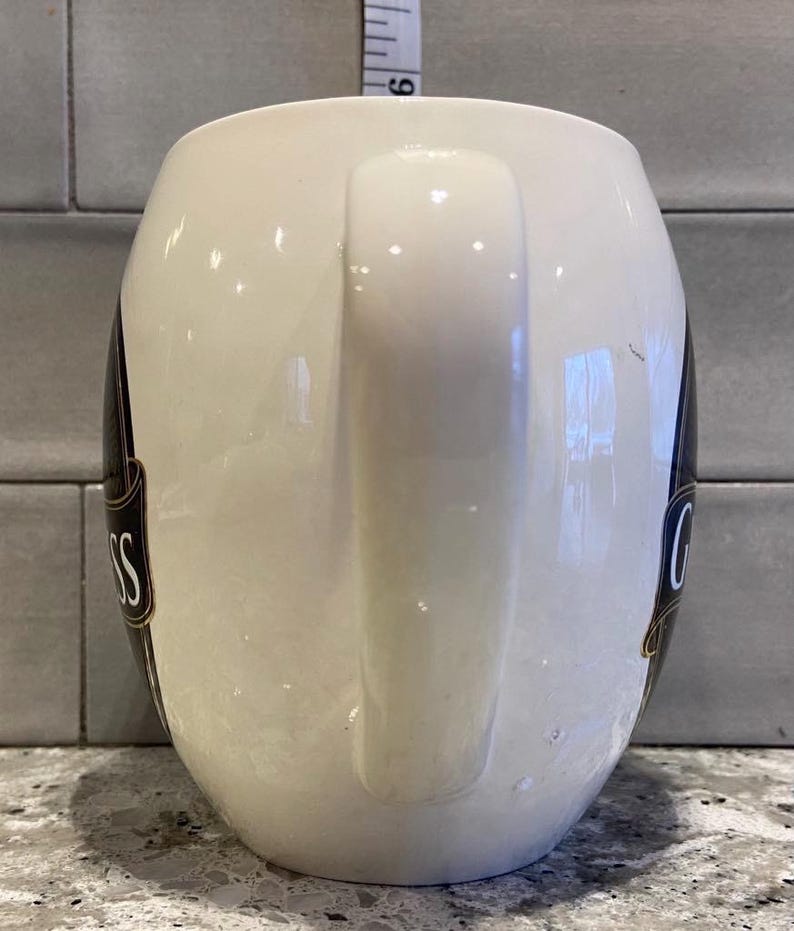 May include: White ceramic mug with a black and gold Guinness logo. The mug has a large handle.