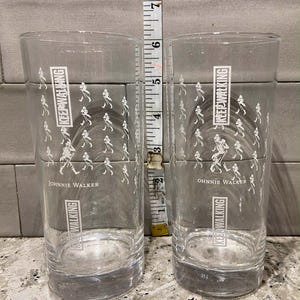 May include: Two clear, cylindrical drinking glasses with the text "KEEP WALKING" and "JOHNNIE WALKER" printed in white. The glasses are approximately 6 inches tall. A white and silver measuring tape is positioned between the glasses.