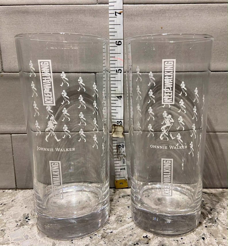 May include: Two clear, cylindrical drinking glasses with the text "KEEP WALKING" and "JOHNNIE WALKER" printed in white. The glasses are approximately 6 inches tall. A white and silver measuring tape is positioned between the glasses.