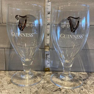 May include: Two clear Guinness beer glasses, each with the Guinness logo. The glasses are about 15 cm tall. They have a stemmed base and a tulip-shaped bowl, designed for enjoying stout.