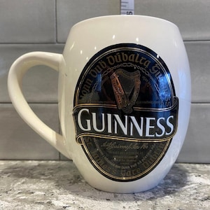 May include: White ceramic mug with a black and gold Guinness logo. The logo features the text "Guinness", "St. James's Gate Dublin", and "Arthur Guinness Son & Co. Ltd. Dublin" in gold lettering. The logo also features a harp and the text "Arthuir Guinness Son & Co. Ltd. Dublin" in black lettering.