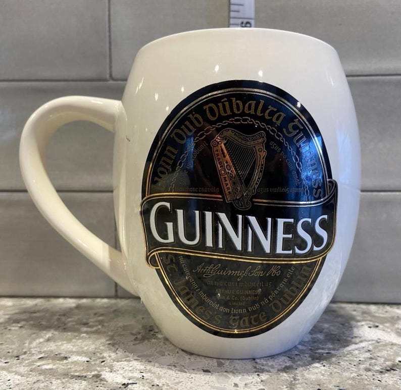 May include: White ceramic mug with a black and gold Guinness logo. The logo features the text "Guinness", "St. James's Gate Dublin", and "Arthur Guinness Son & Co. Ltd. Dublin" in gold lettering. The logo also features a harp and the text "Arthuir Guinness Son & Co. Ltd. Dublin" in black lettering.