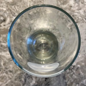 May include: A clear, empty drinking glass with a rounded shape. The glass has a thick rim and sits on a speckled gray surface. The glass is transparent, allowing a view through its interior.