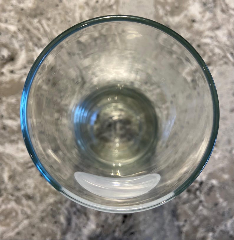 May include: A clear, empty drinking glass with a rounded shape. The glass has a thick rim and sits on a speckled gray surface. The glass is transparent, allowing a view through its interior.