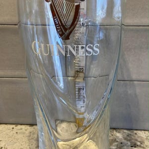 Op de afbeelding: Een helder Guinness pintglas met het logo en de tekst van het merk. Het glas heeft een gebogen ontwerp en is ongeveer 18 cm hoog. Een meetlint zit in het glas.