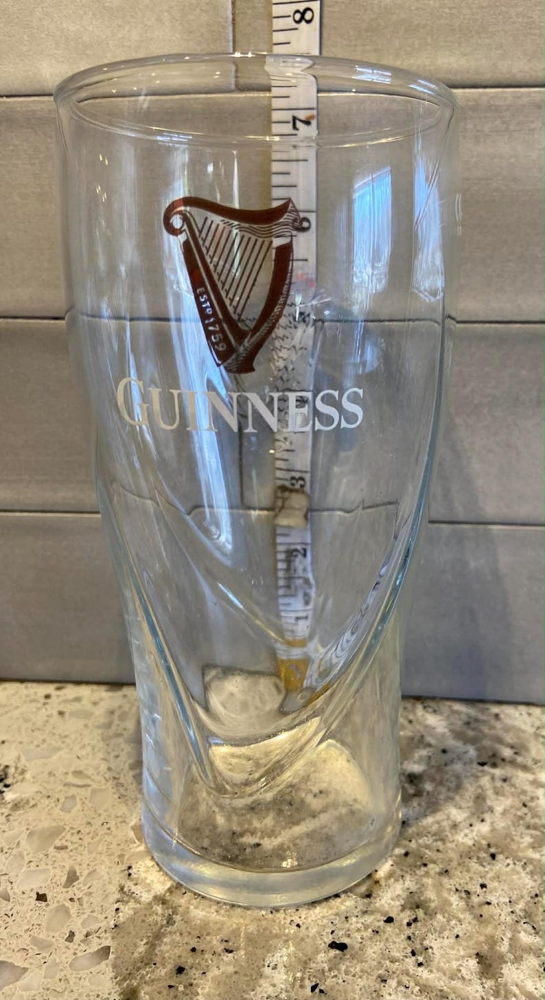 May include: A clear Guinness pint glass with the brand's logo and text. The glass has a curved design and is approximately 7 inches tall. A measuring tape is inside the glass.