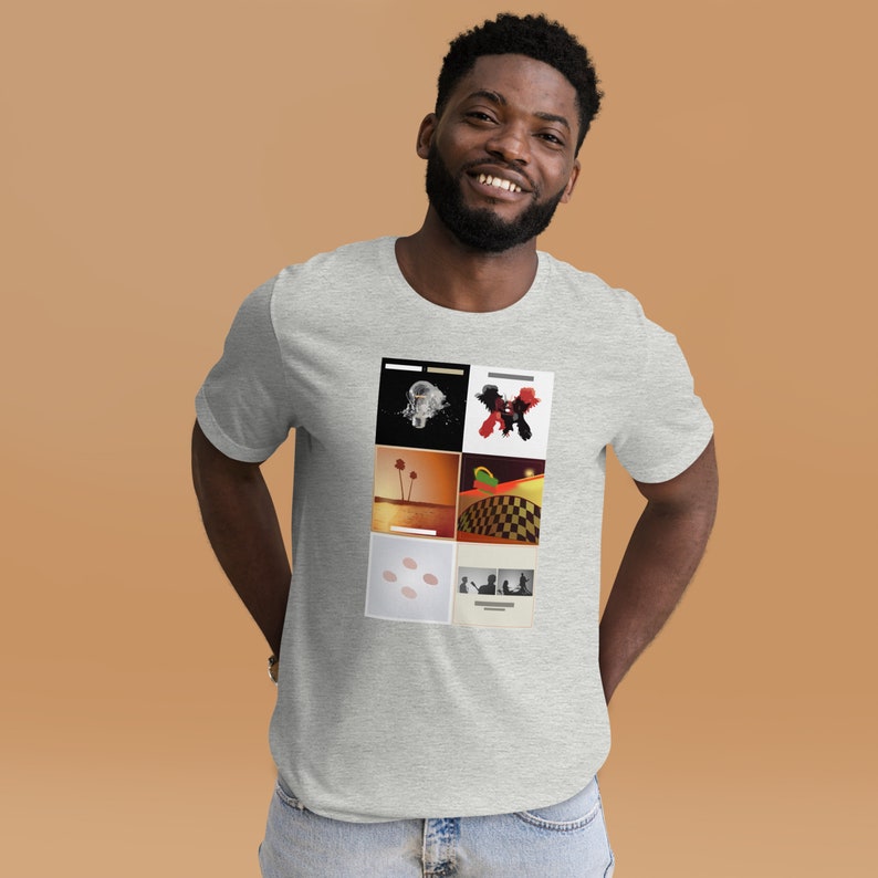 Puede incluir: Una camiseta gris con un collage de seis portadas de &aacute;lbumes diferentes. Las portadas de los &aacute;lbumes presentan una variedad de colores y dise&ntilde;os, incluyendo una imagen en blanco y negro de una persona con un traje espacial, un dise&ntilde;o abstracto rojo y negro, un paisaje des&eacute;rtico con palmeras, un patr&oacute;n a cuadros, un primer plano de un objeto rosa y blanco, y una imagen en blanco y negro de dos personas.