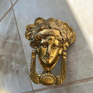 May include: A gold-toned door knocker with a detailed design of a woman's face. The knocker has a ring-shaped handle and decorative wings.