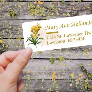 Puede incluir: Una etiqueta de dirección blanca con un diseño floral con flores amarillas y hojas verdes. La etiqueta tiene el texto "Mary Ann Hollander", "1234 St. Lawrence Hwy." y "Lewiston, Id 23456" impreso en dorado.