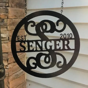 May include: A circular black metal sign hanging from a black metal post by two chains. The sign features a large stylized letter S in the center, the text SENGER across the middle, and the text EST. 2010 on either side of the name.