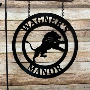 May include: Black metal garden sign with the text "Wagner's Manor" in a circular design. A lion silhouette is centered within the circle. The sign hangs from a black metal stake with a curved top.