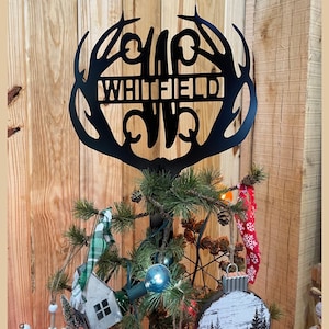 May include: Black metal tree topper with the name "WHITFIELD" cut out, framed by stylized antlers. The topper is placed on a decorated Christmas tree with ornaments, lights, and pinecones. The background is a wooden wall.